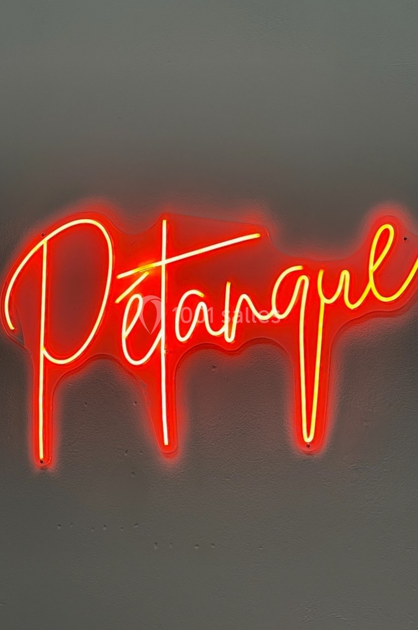 Enseigne néon rouge affichant le mot ’Pétanque’ sur un mur gris uni.