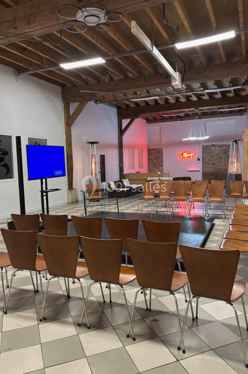 Salle avec des chaises disposées en cercle autour d'une petite scène, écran allumé et éclairage moderne.