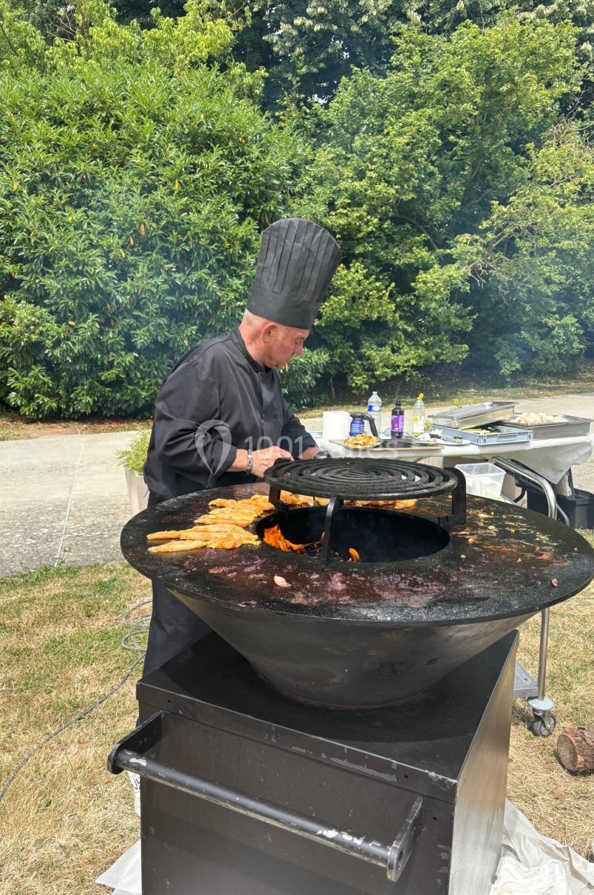 Un chef en tenue noire cuisine des aliments sur une grande plancha ronde en extérieur, entouré de verdure.