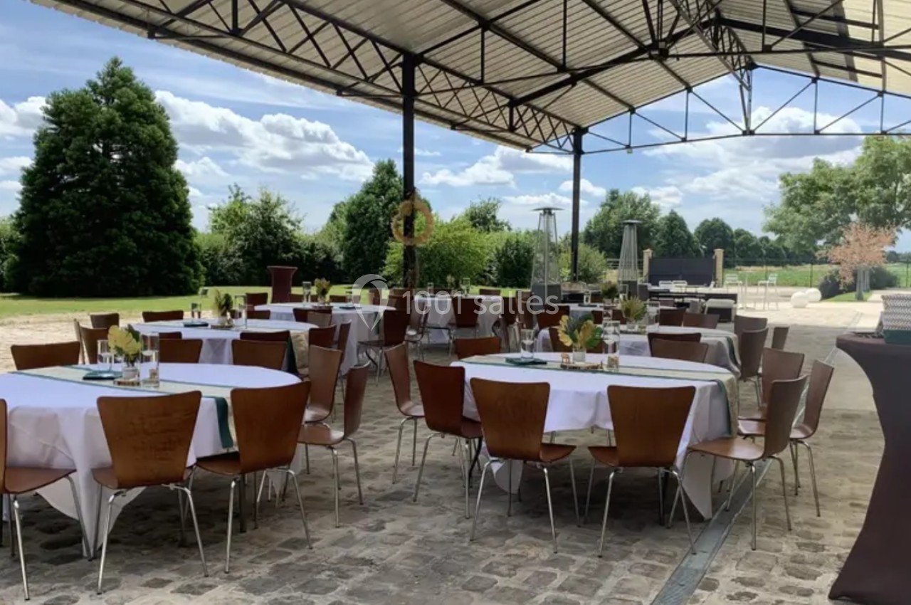Tables dressées avec nappes blanches et chaises en bois sous une structure couverte, entourées de verdure et ciel dégagé.