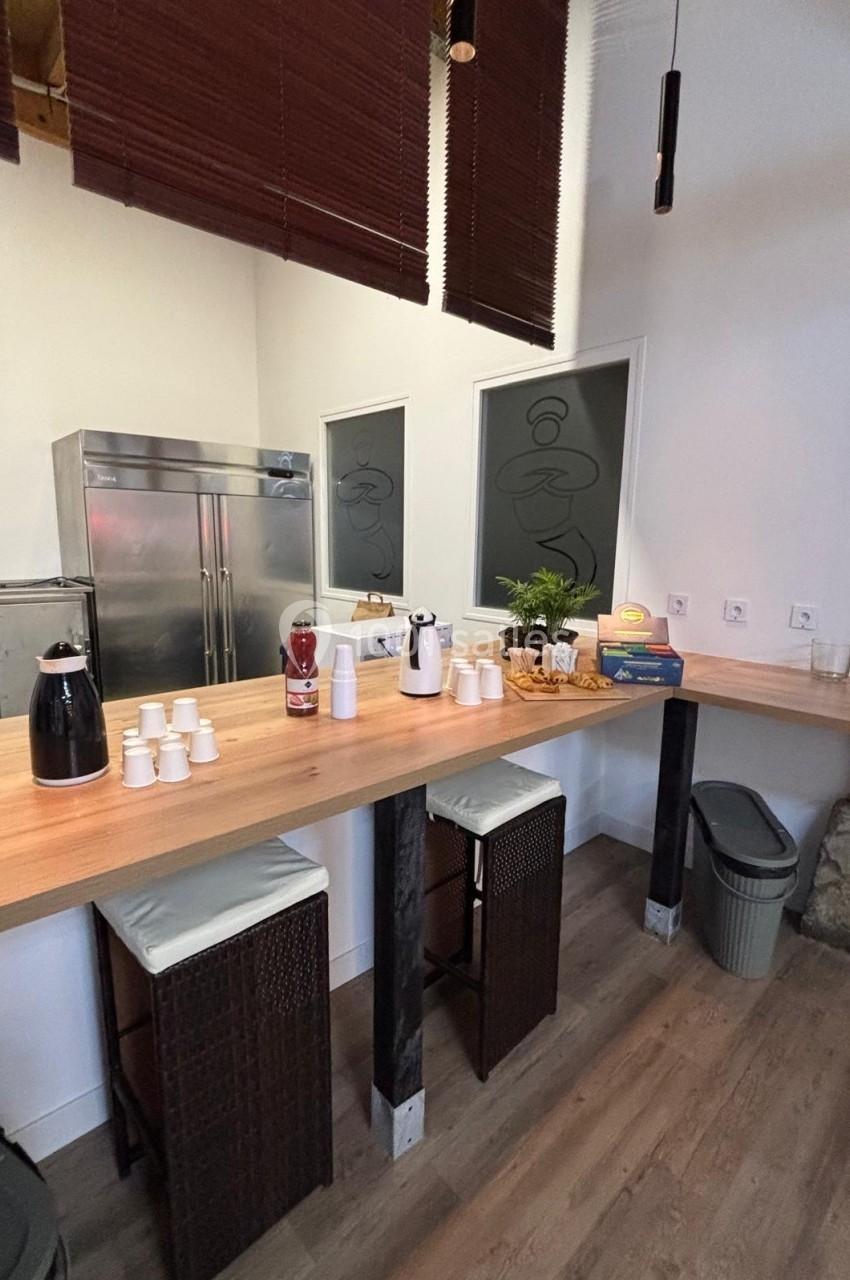 Comptoir en bois avec cafetière, gobelets, plantes et tabourets, situé dans un espace lumineux avec un sol en bois.