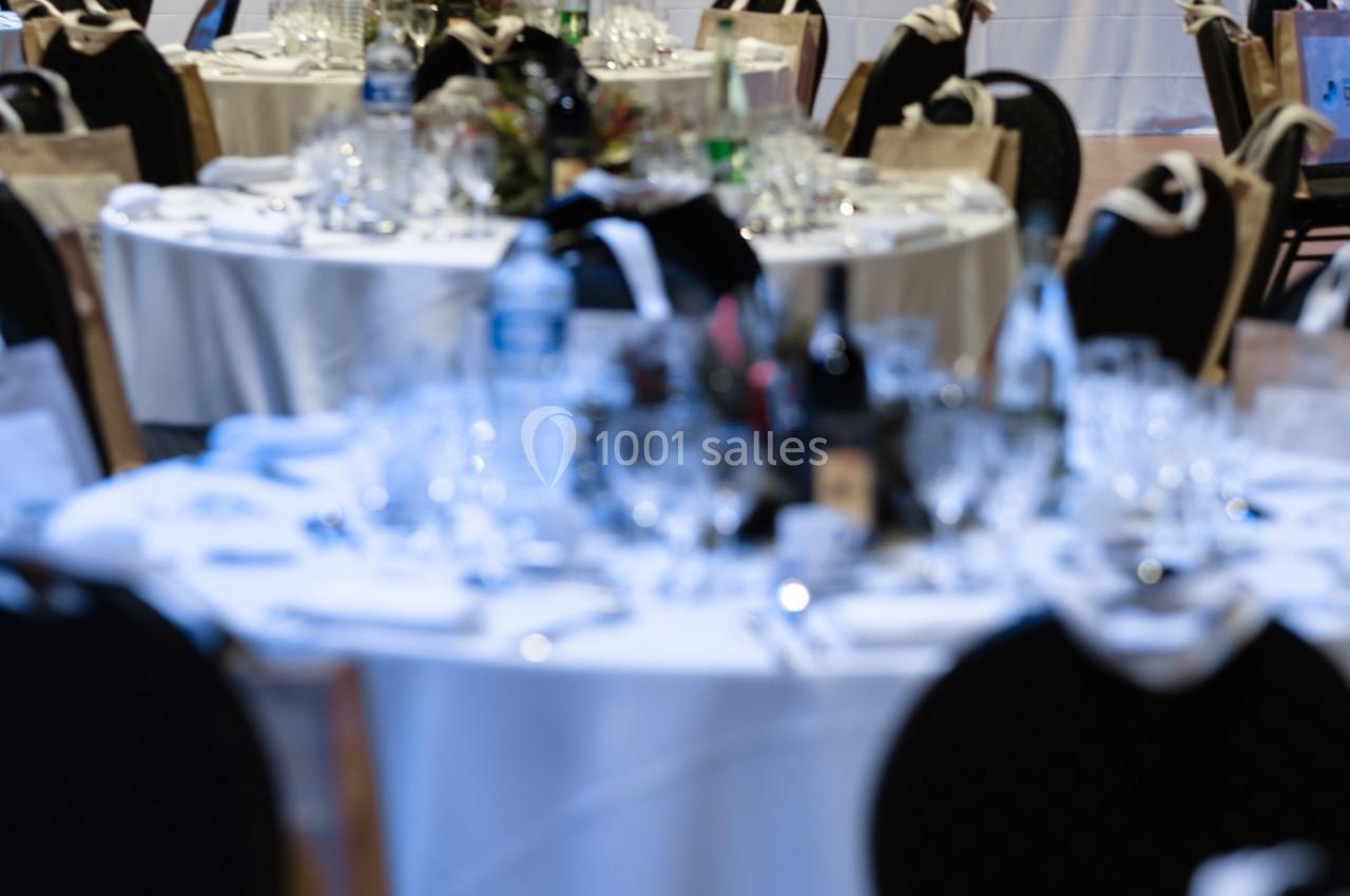 Tables rondes dressées avec nappes blanches, verres et bouteilles, devant un buffet avec verres empilés et seaux à glace.