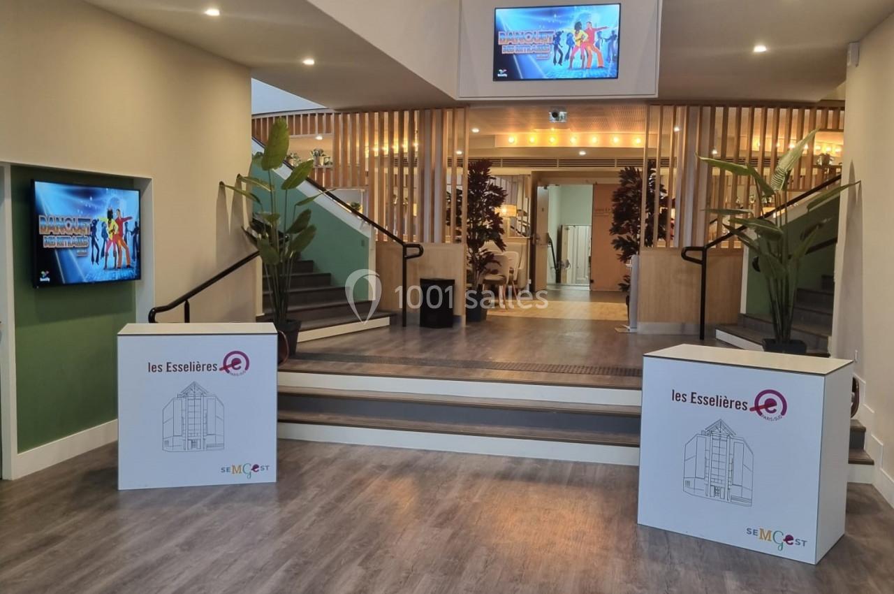 Entrée d'un bâtiment avec escalier central, écrans d'affichage, plantes décoratives et sol en parquet.