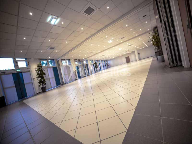 Salle spacieuse et lumineuse avec un sol carrelé, des murs vitrés et un éclairage au plafond.