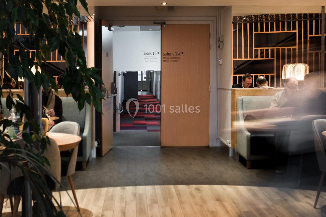 Vue d'un espace intérieur avec des tables, des fauteuils et une porte ouverte menant à un couloir éclairé.