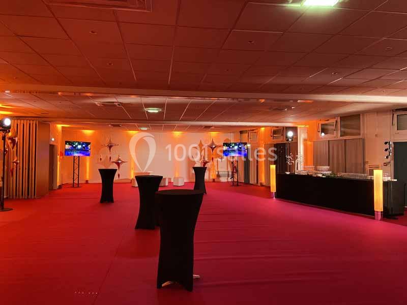 Salle de réception avec éclairage orange, tables hautes noires et écrans disposés sur les côtés.