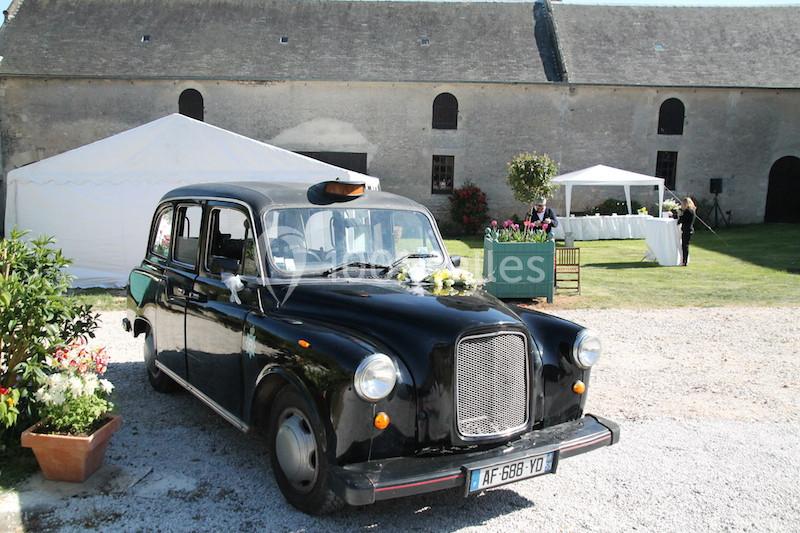 Taxi noir ancien garé devant une grange en pierre avec des tentes blanches et un jardin fleuri en arrière-plan.