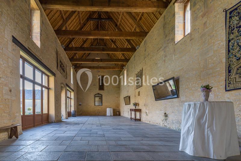 Grande salle en pierre avec plafond en bois, fenêtres vitrées, tapisseries murales et table recouverte d'une nappe blanche.