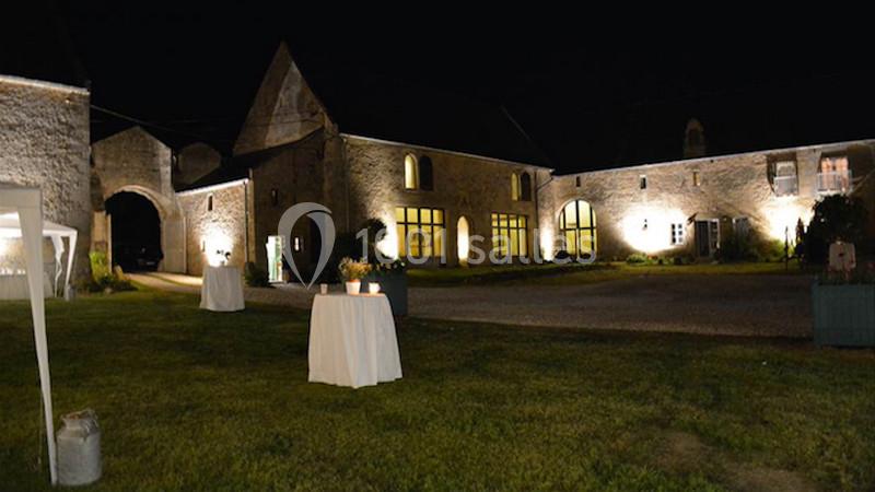 Cour d'une bâtisse en pierre éclairée la nuit, avec tables dressées et pelouse au premier plan.