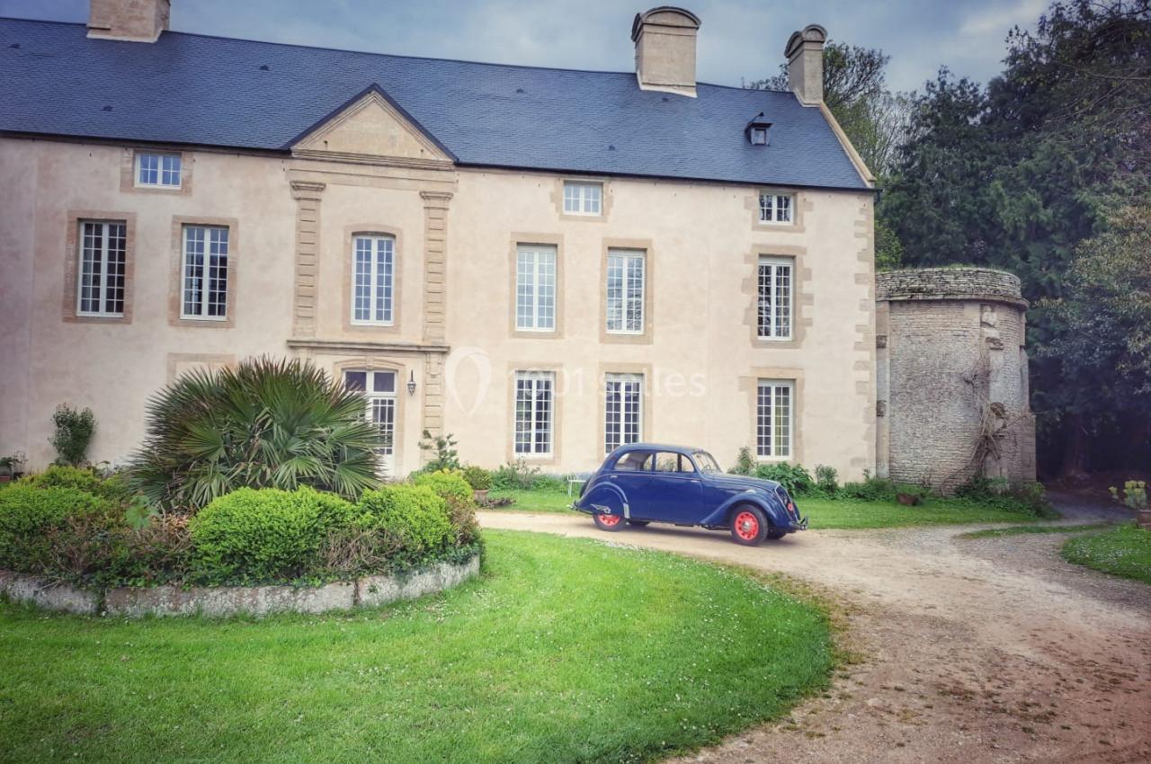 Façade d'une grande maison en pierre avec une voiture ancienne bleue garée devant, entourée de verdure.