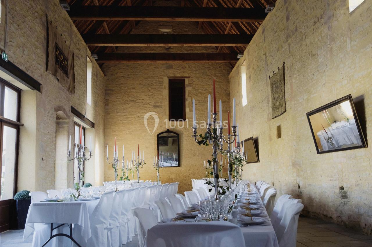 Grande salle en pierre avec tables dressées pour un banquet, ornées de chandeliers et de nappes blanches.