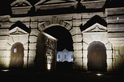 Vue nocturne d'une porte monumentale en pierre éclairée, avec un bâtiment blanc visible en arrière-plan.