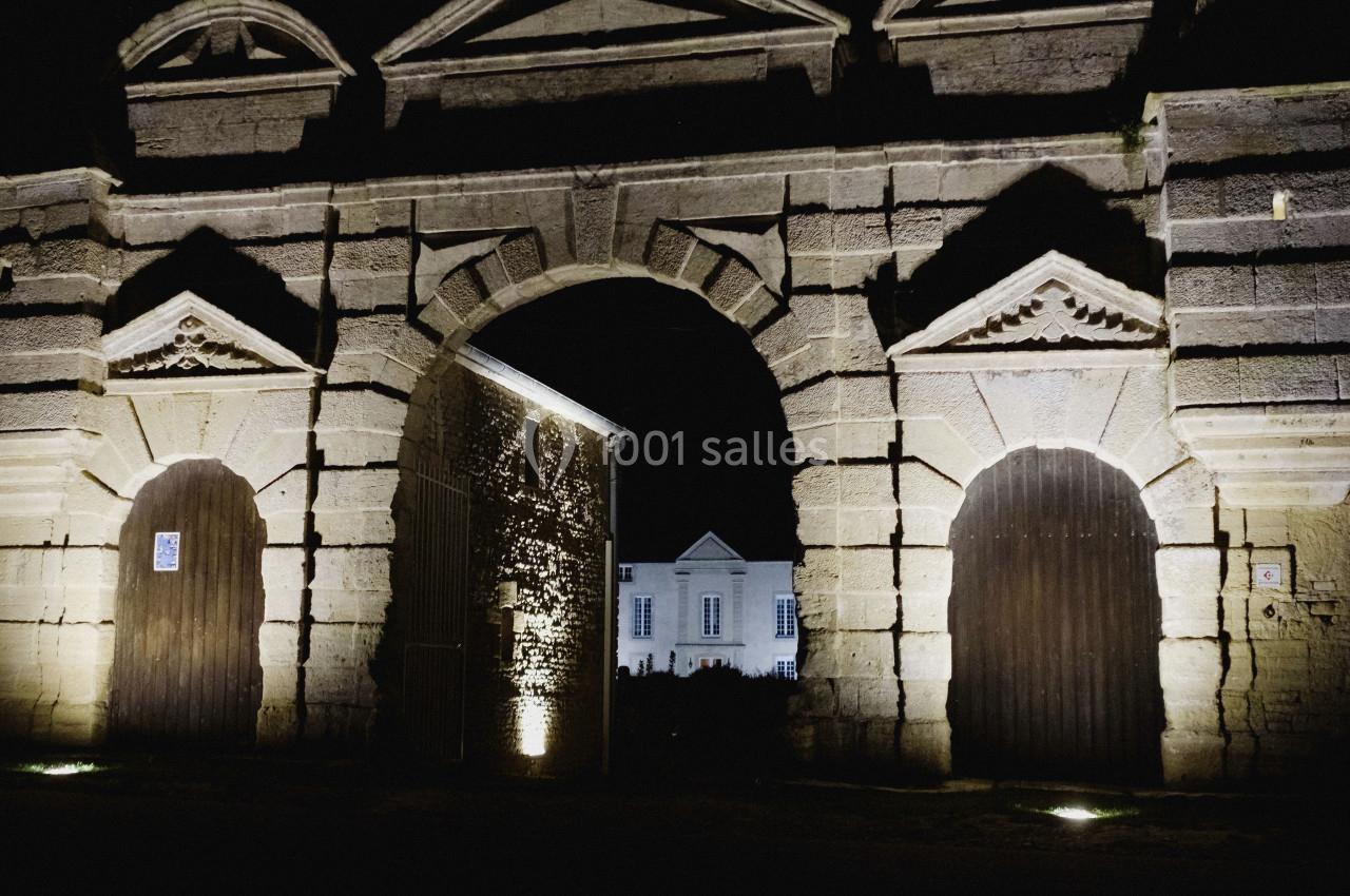 Vue nocturne d'une porte monumentale en pierre éclairée, avec un bâtiment blanc visible en arrière-plan.