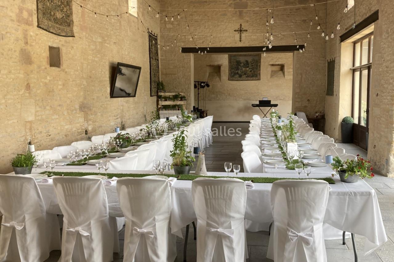 Salle en pierre avec tables dressées en U, nappes blanches, chaises couvertes, décorations végétales et guirlandes…