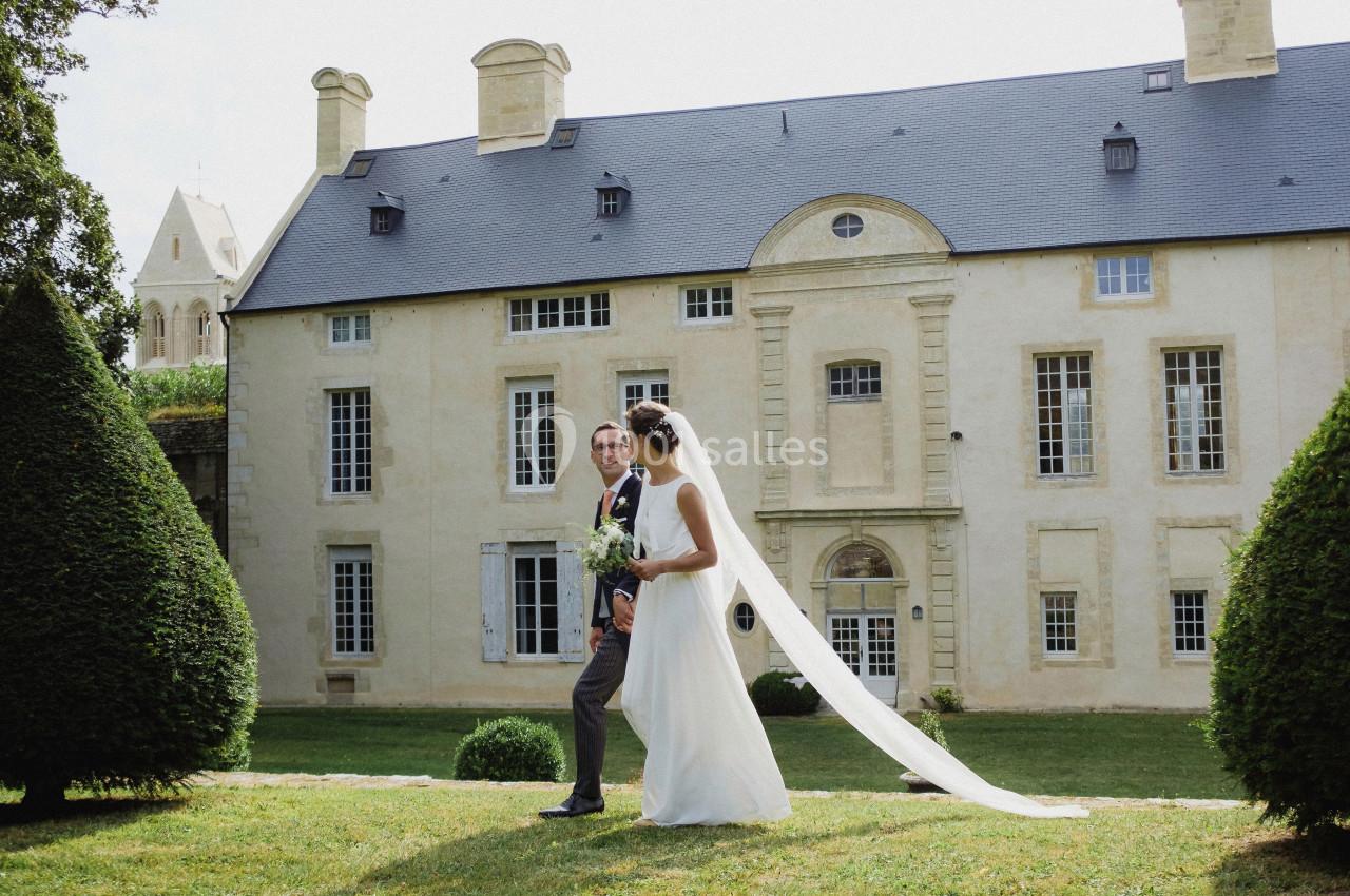 Un couple de mariés marche sur une pelouse devant un bâtiment historique en pierre, par une journée ensoleillée.