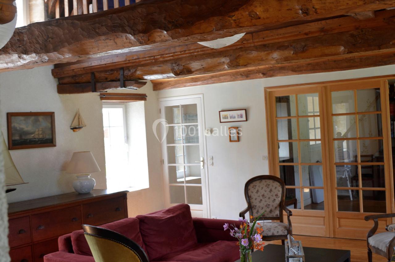 Salon chaleureux avec poutres apparentes, canapé rouge, fauteuils, table basse et lumière naturelle entrant par les fenêtres.