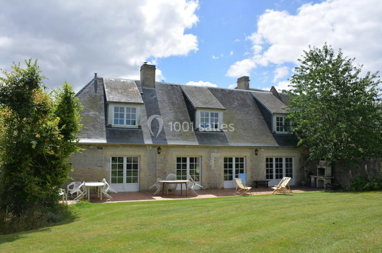 Maison en pierre avec toit en ardoise, grandes fenêtres, terrasse aménagée et jardin verdoyant sous un ciel partiellement…