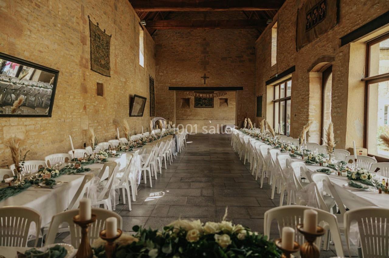 Grande salle en pierre aménagée pour un banquet, avec longues tables décorées de fleurs et chaises blanches.