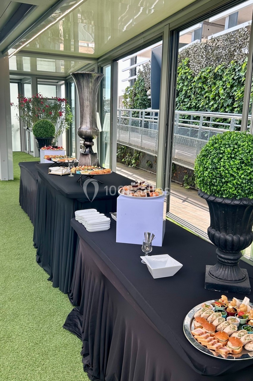 Buffet avec plateaux de nourriture variée disposés sur des tables drapées de noir dans un espace lumineux avec vue…