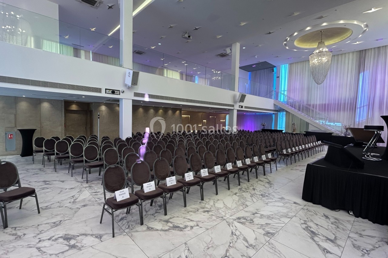 Salle de conférence moderne avec rangées de chaises, scène surélevée et éclairage coloré.
