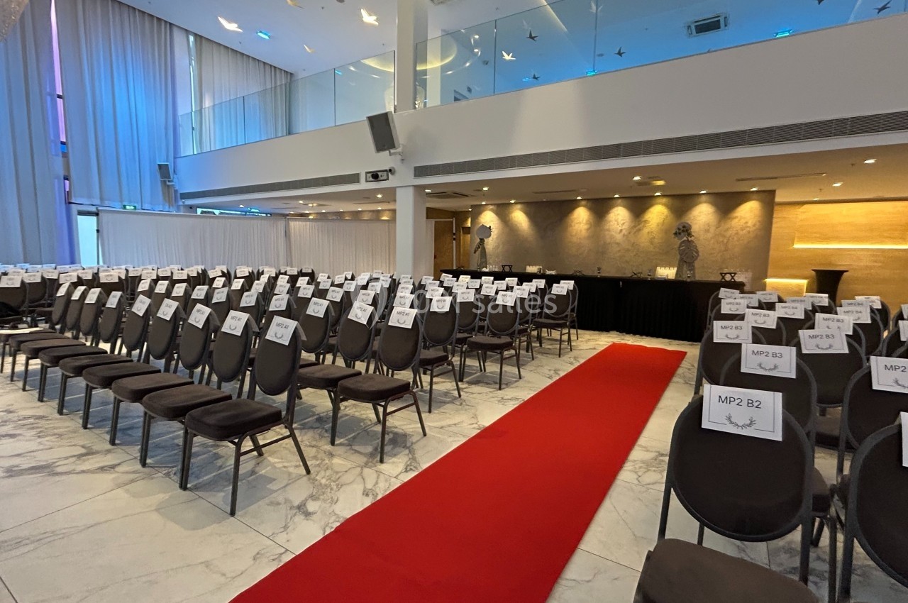 Salle de conférence avec rangées de chaises noires alignées, tapis rouge central et étiquettes de placement sur les sièges.