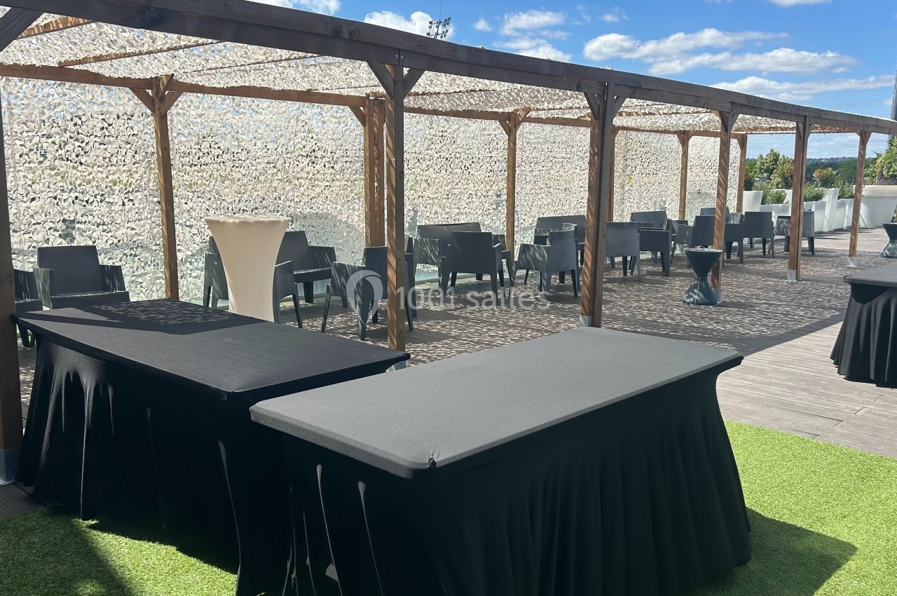 Terrasse aménagée avec tables drapées de noir, chaises et pergola en bois sous un ciel ensoleillé.