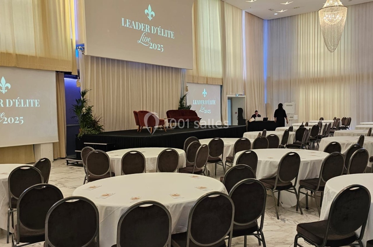 Salle de conférence lumineuse avec tables rondes, scène équipée de fauteuils et écrans affichant ’Leader d'Élite Live 2025’.