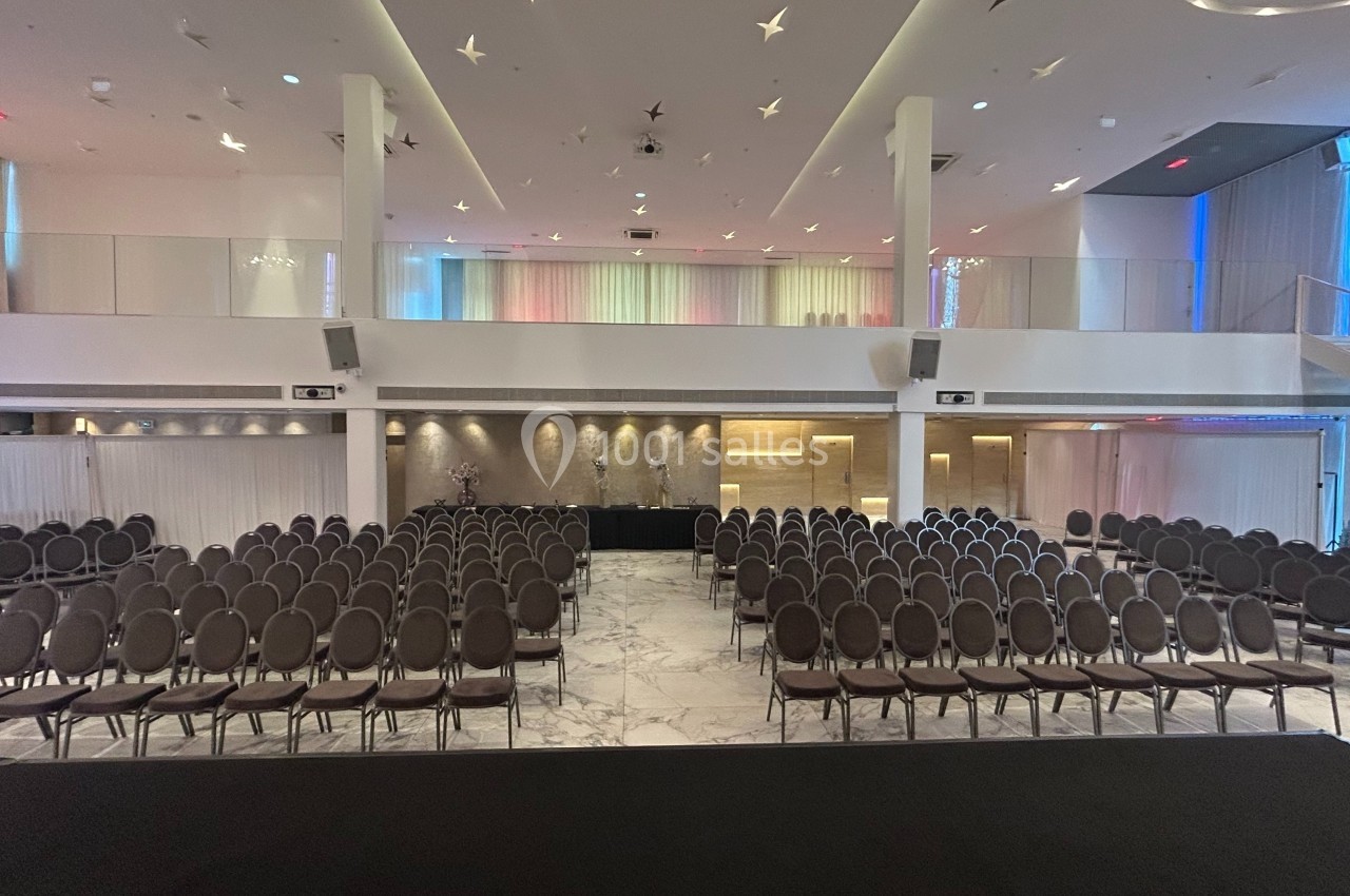 Salle de conférence moderne avec rangées de chaises alignées, scène noire au premier plan et éclairage tamisé.