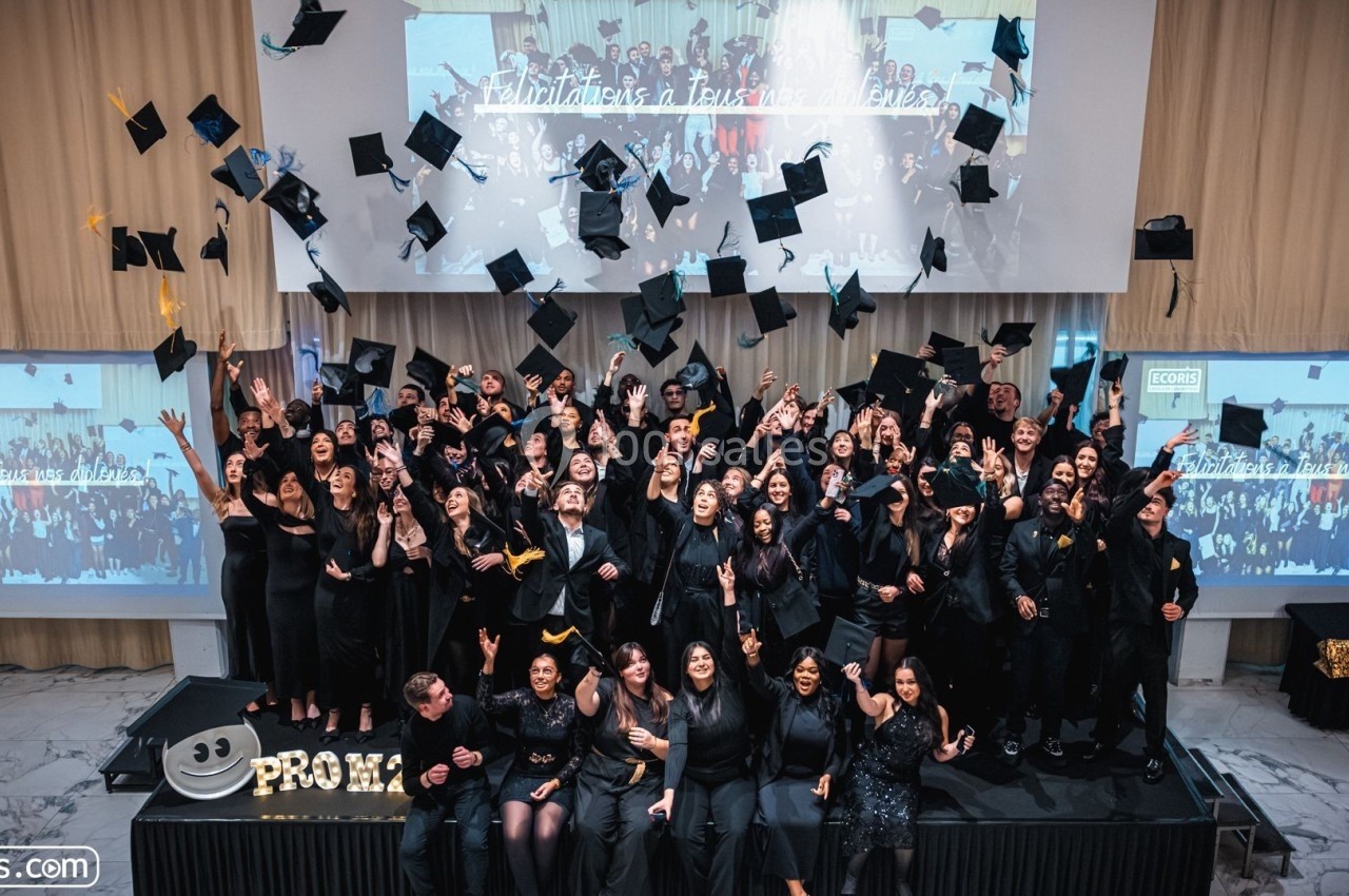 Des diplômés en tenue de cérémonie lancent leurs toques en l'air devant un écran affichant une photo de groupe.