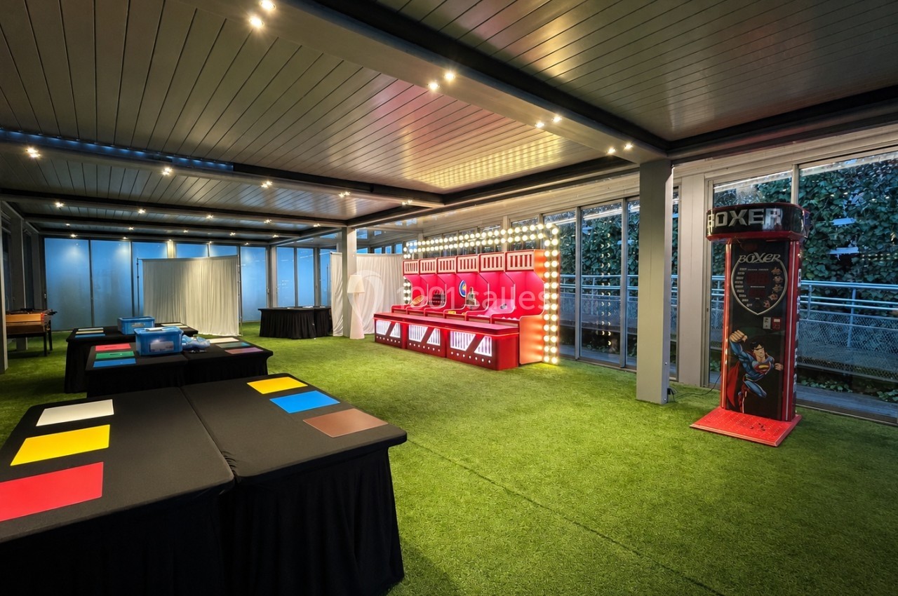 Salle de jeux intérieure avec stands colorés, machine de boxe et tables sur gazon synthétique, éclairage moderne.