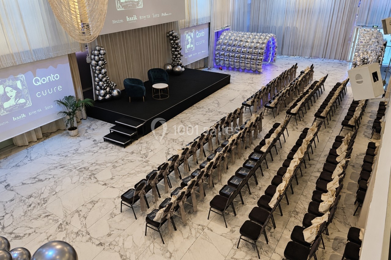 Salle de conférence moderne avec rangées de chaises, scène décorée et écrans projetant des visuels.