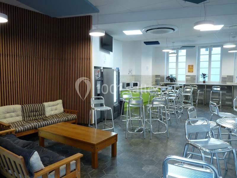 Salle de pause lumineuse avec tables, chaises en métal, canapé, table basse et coin cuisine équipé.