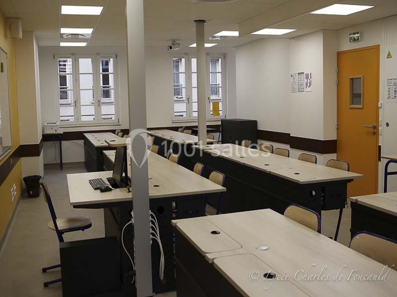 Salle de classe vide avec des bureaux alignés, des chaises, un ordinateur et des fenêtres donnant sur l'extérieur.