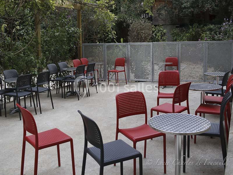 Terrasse extérieure avec des tables rondes et des chaises rouges et noires, entourée de végétation et de barrières…