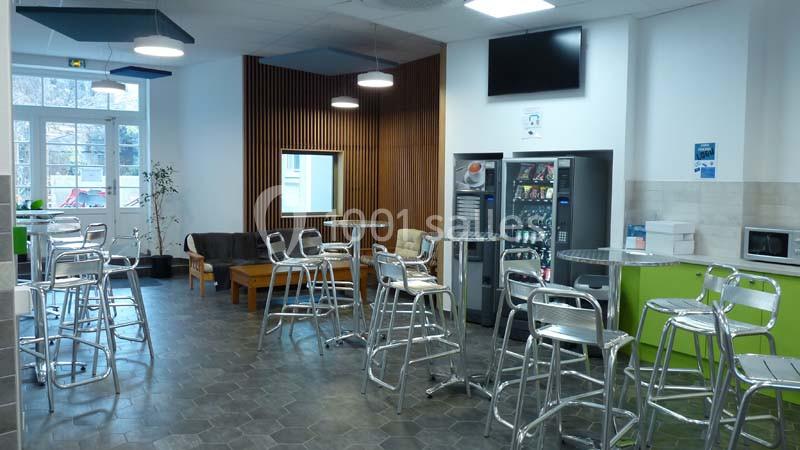 Salle de pause lumineuse avec tables, chaises en métal, distributeurs automatiques et coin détente avec canapé.