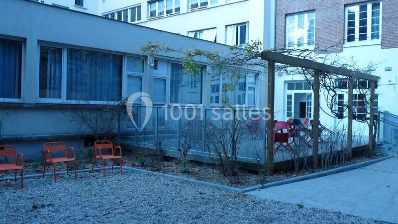 Cour intérieure avec sol en gravier, chaises orange, terrasse surélevée vitrée et bâtiments en arrière-plan.