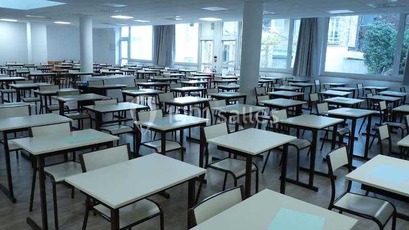 Salle de classe vide avec des rangées de tables et chaises alignées, éclairée par des fenêtres donnant sur l'extérieur.