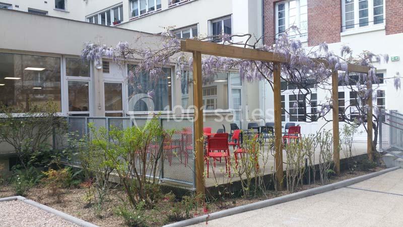 Terrasse extérieure avec pergola en bois, glycines en fleurs, chaises rouges et bâtiment en arrière-plan.