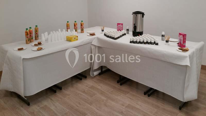 Tables dressées avec boissons, gobelets, biscuits et thermos dans une salle sobre au sol en bois.