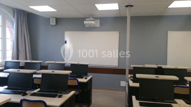 Salle de classe équipée d'ordinateurs, de bureaux alignés, d'un tableau blanc et d'un projecteur au plafond.