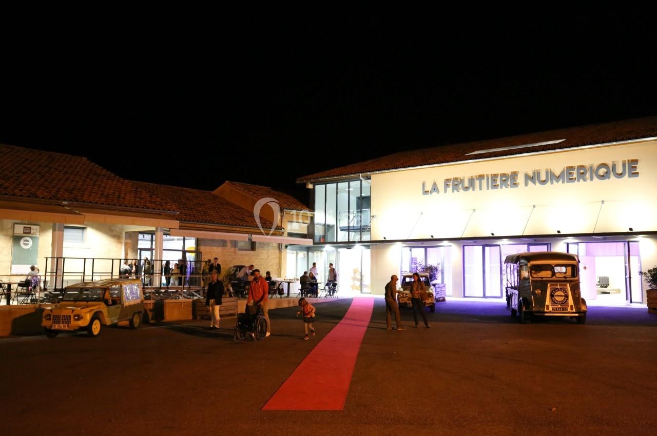 Entrée éclairée de nuit d'un bâtiment nommé ’La Fruitière Numérique’, avec des visiteurs et des véhicules anciens.
