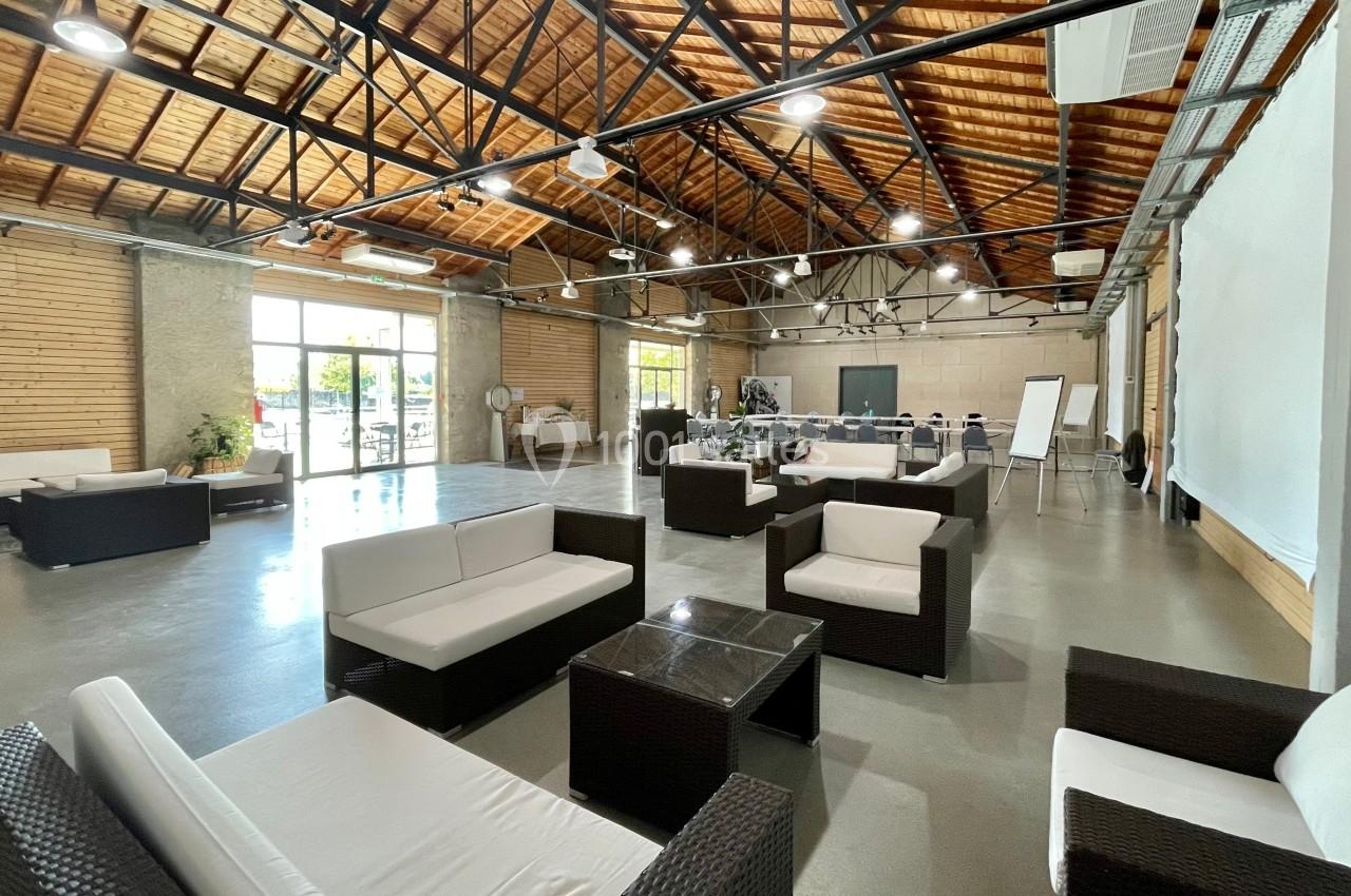 Salle spacieuse avec mobilier en rotin, grandes baies vitrées, plafond en bois et structure métallique apparente.