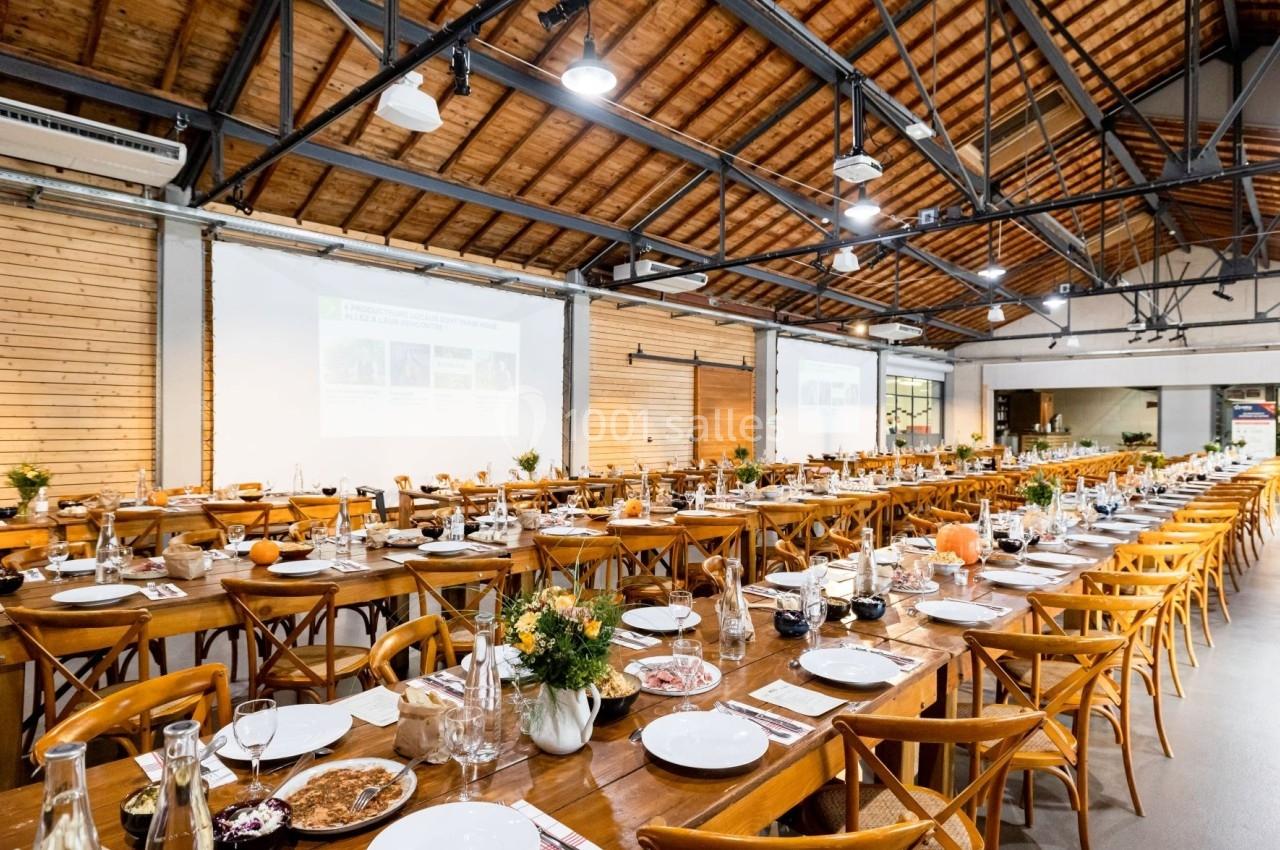 Salle de réception avec de longues tables en bois dressées pour un repas, sous un plafond en poutres apparentes.