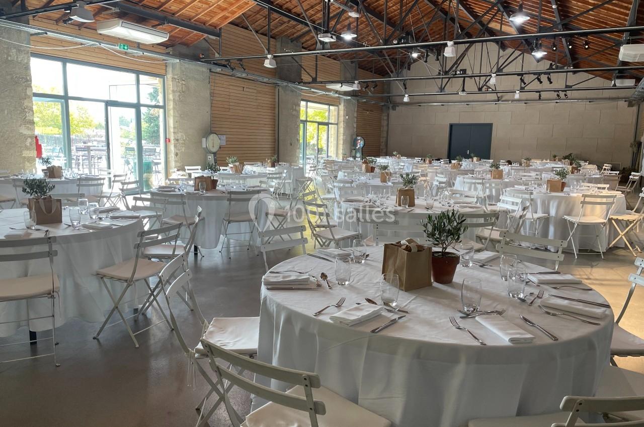 Salle de réception lumineuse avec tables rondes dressées de nappes blanches, couverts et décorations simples.