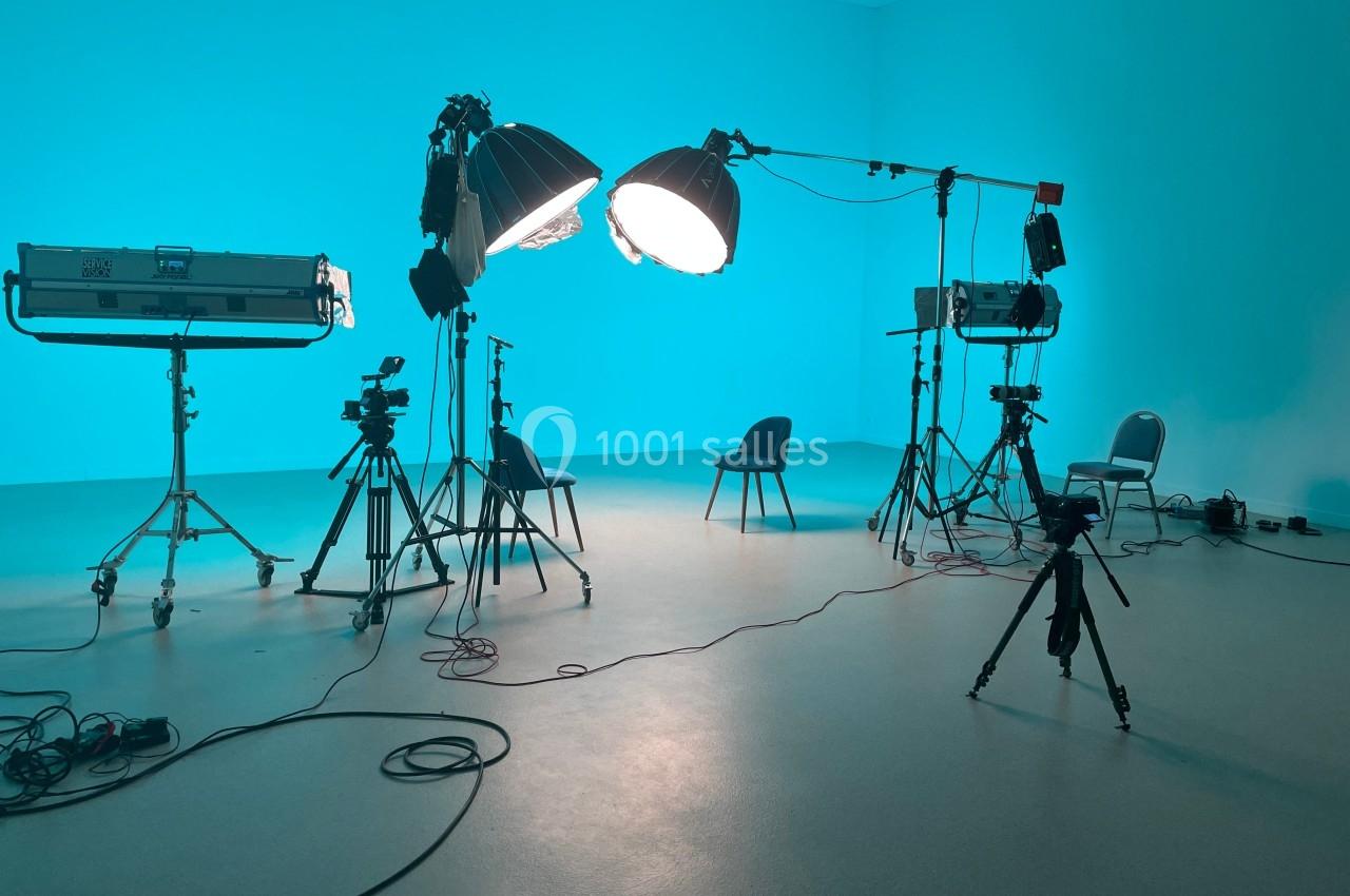 Studio photo avec éclairages, trépieds, caméras et chaises dans une pièce aux murs bleus.