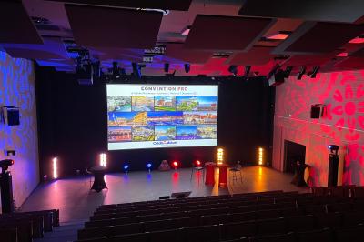 Salle de réunion avec tables et chaises disposées en U, grand écran blanc, plantes et buffet en arrière-plan.
