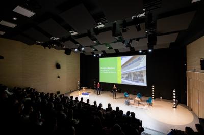 Salle de réunion avec tables et chaises disposées en U, grand écran blanc, plantes et buffet en arrière-plan.