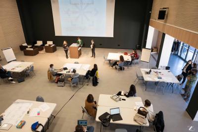 Salle de réunion avec tables et chaises disposées en U, grand écran blanc, plantes et buffet en arrière-plan.