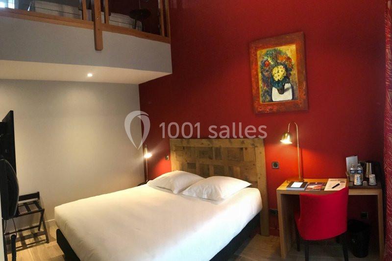 Chambre avec lit double, mur rouge décoré d'un tableau, bureau avec chaise rouge et éclairage chaleureux.