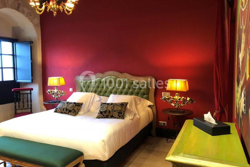 Chambre avec lit double, tête de lit capitonnée verte, murs rouges, éclairage chaleureux et mobilier classique.